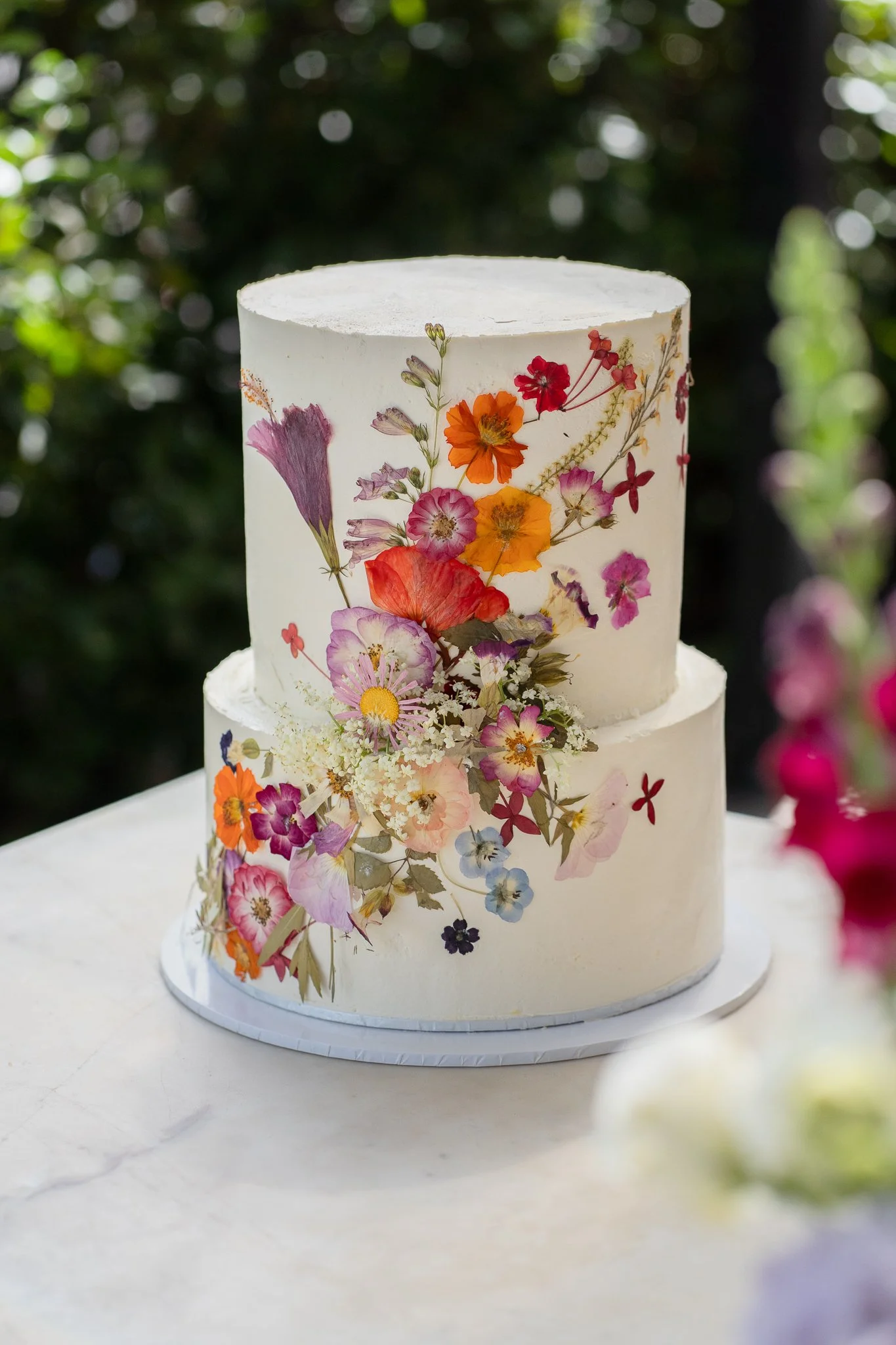 A two-tiered white wedding cake decorated with caramelized fruit and edible flowers on a white table outdoors.