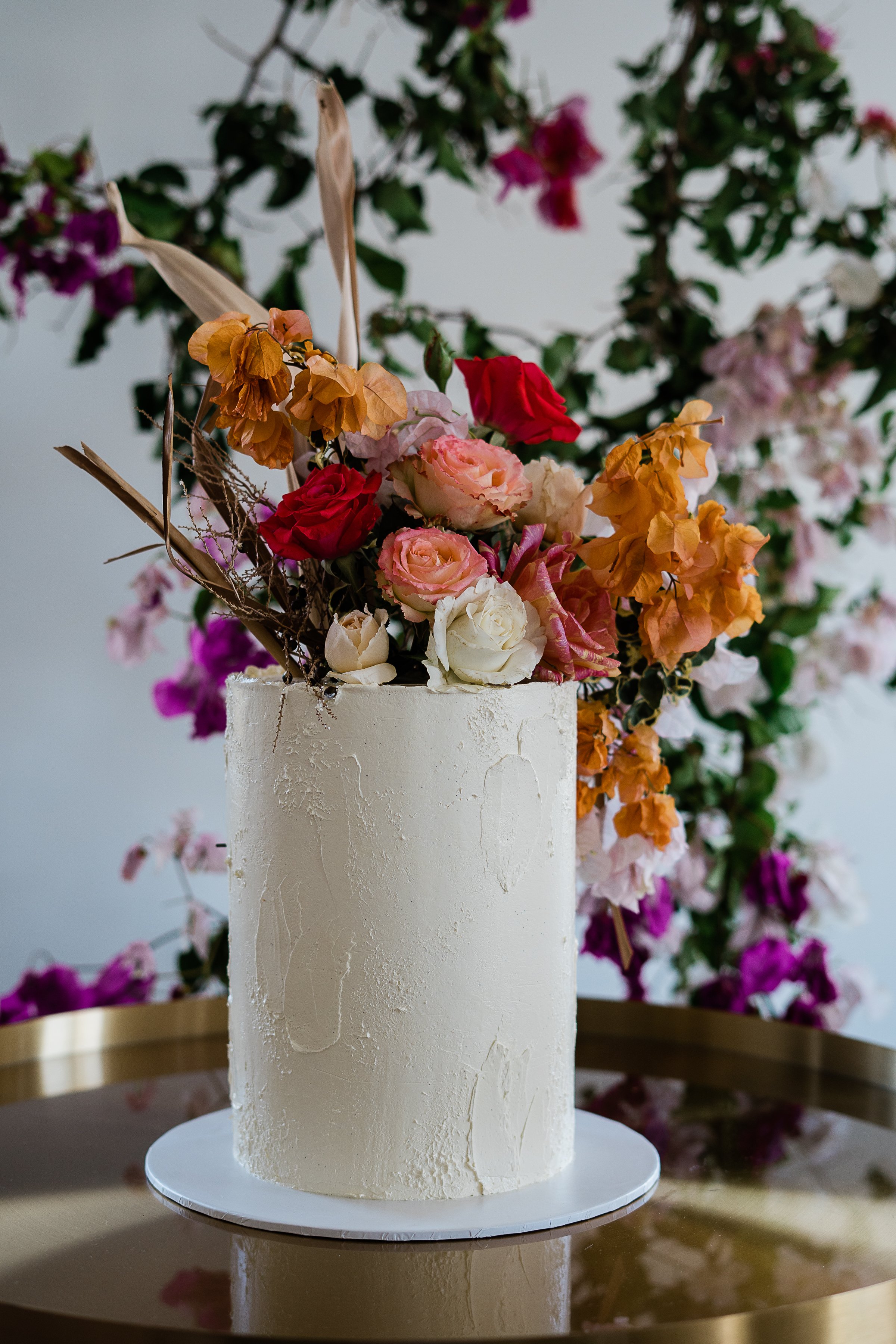 A white cake adorned with a floral arrangement on top, including pink, red, yellow, and white roses, with a background of pink and purple flowers and green leaves.