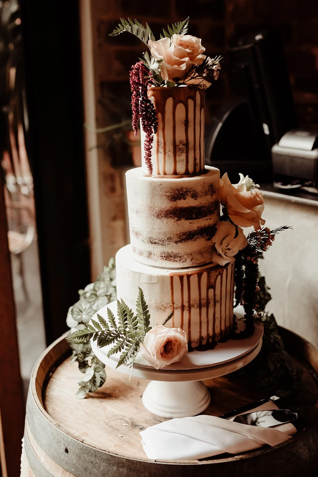 A three-tiered wedding cake decorated with fresh flowers and greenery, featuring a semi-naked style with minimal frosting and caramel drip accents, placed on a wooden barrel with ribbons and flowers