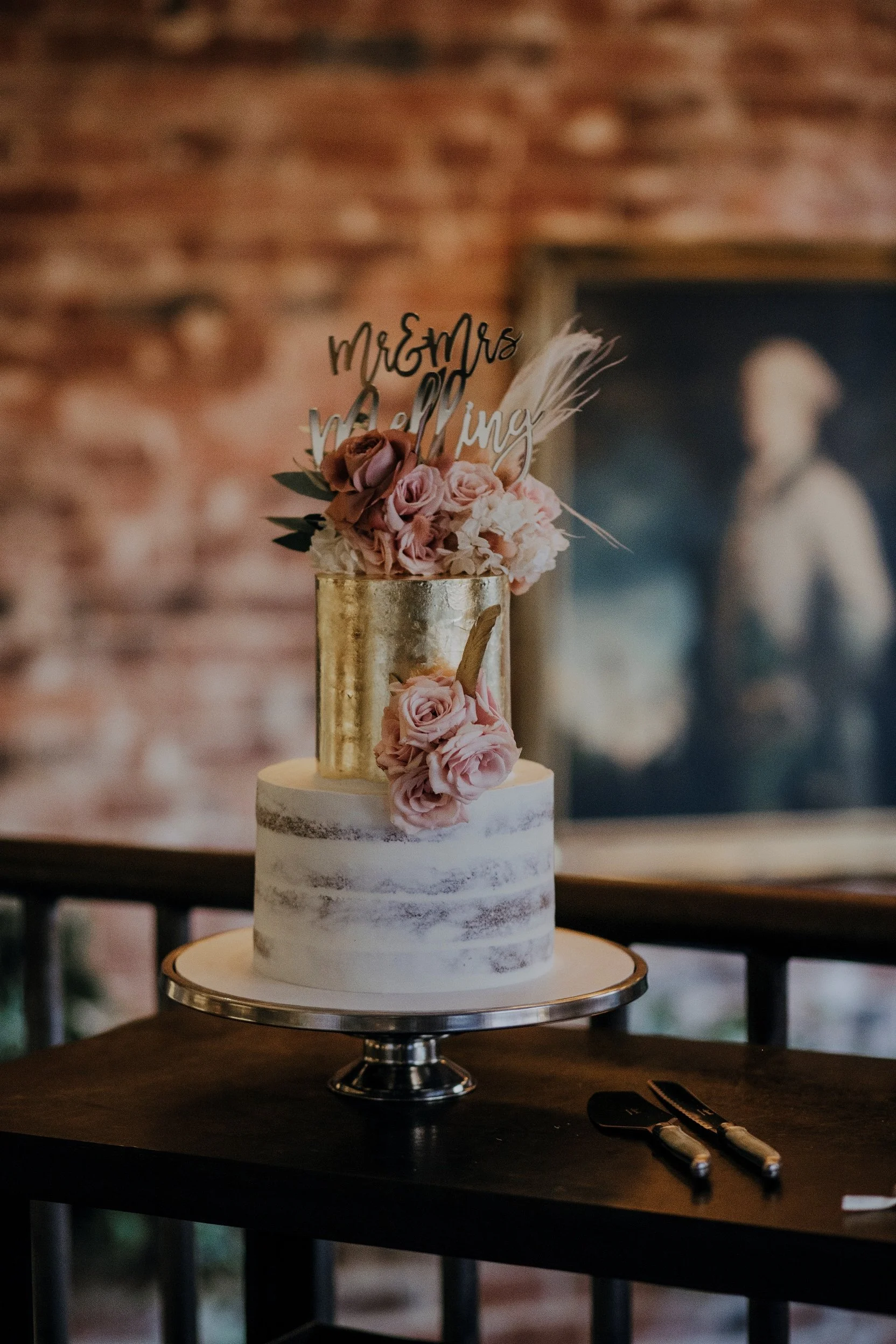 A wedding cake with a white and gray marble pattern, topped with pink and blush roses, greenery, and a 'Mr & Mrs' cake topper, placed on a round cake stand in front of a brick wall. A blurred bride in a wedding dress is visible in the background.