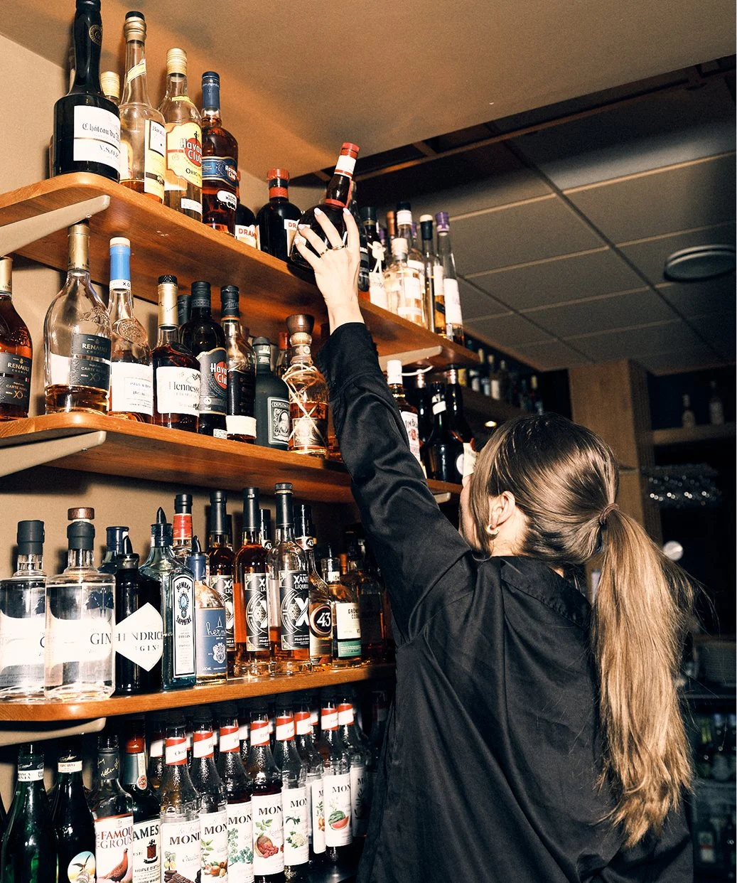 Kvinna som tar ner en flaska från en övre hylla i en bar eller butik med många olika sorts spritflaskor på hyllorna.