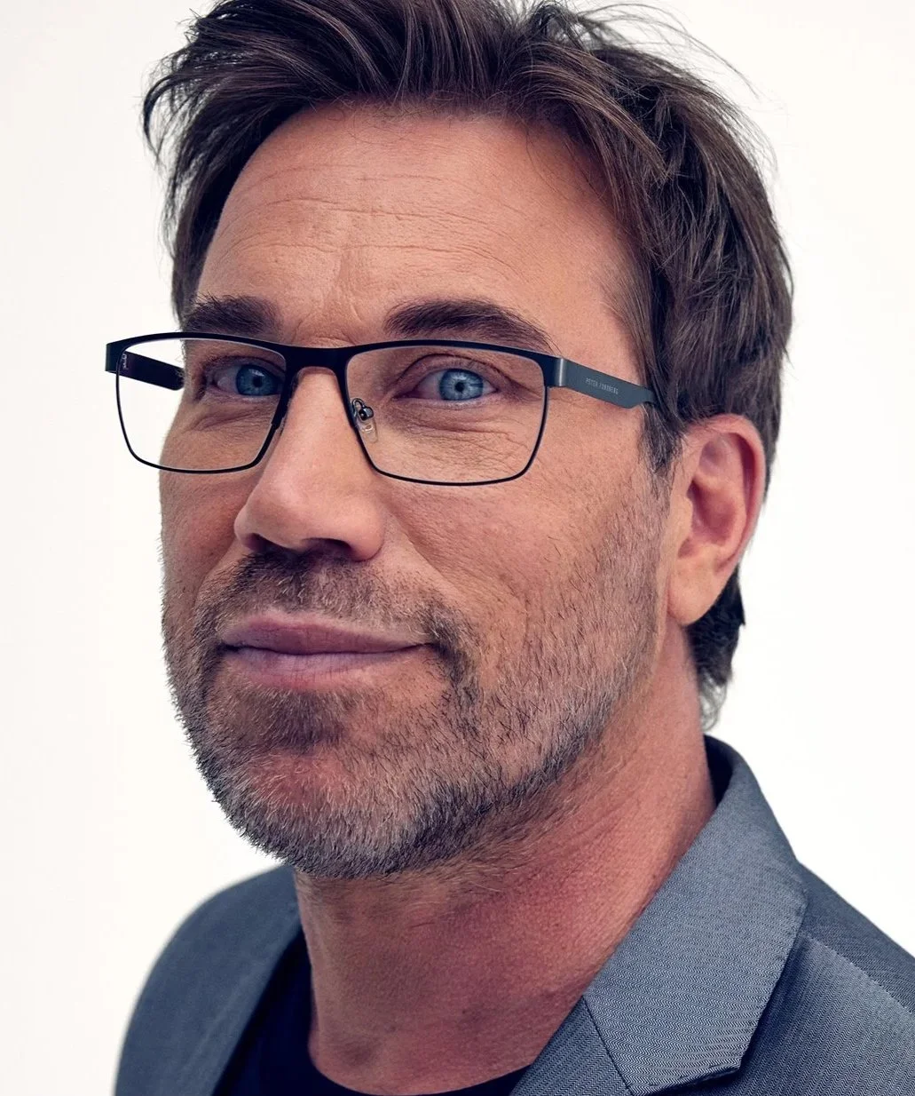 En man med blå ögon, mörkt hår och stubb, bär glasögon och en grå blazer, står mot en vit bakgrund.