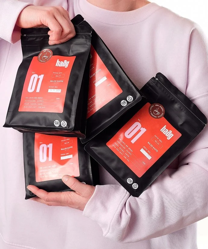 En person som bär en ljusrosa tröja håller i fyra svarta förpackningar av kaffe med röda etiketter.