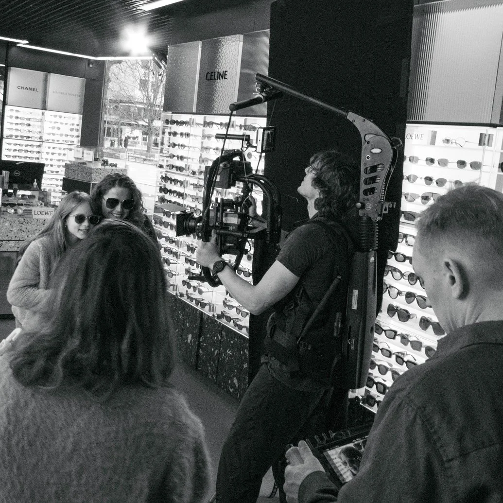 Flera personer vid en fotosession i en optikaffär. Två kvinnor bär solglasögon framför ett bord fullt av olika solglasögon. En man med en kamera med gimbal filmar dem.