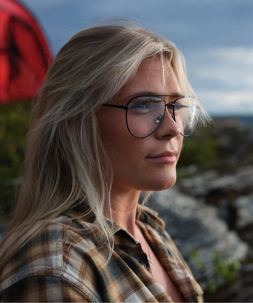 En blond kvinna med glasögon tittar ut mot horisonten på en utomhusplats under molnig himmel, med ett berg eller klippor i bakgrunden.