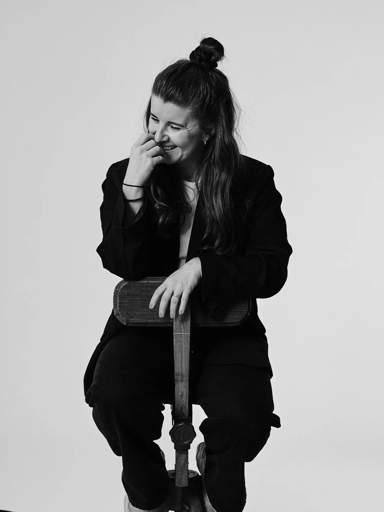 Svartvit bild av en ung kvinna som skrattar och lutar sig mot en skateboard, sittande mot en tom vit bakgrund.