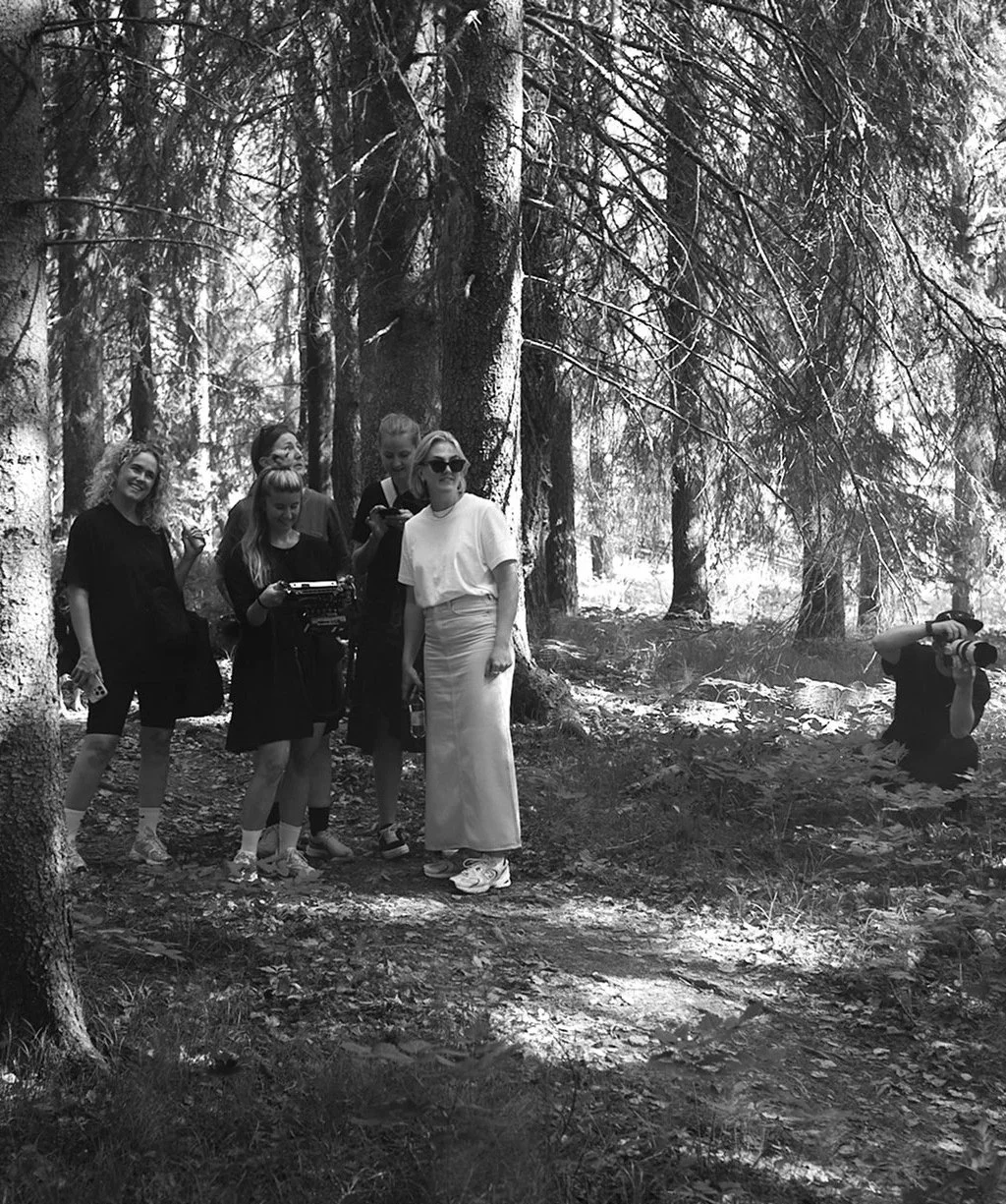 Fem personer, fyra kvinnor och en man, står i en skog. En av kvinnorna bär solglasögon och en annan håller en dryck. En person till vänster håller något i handen. En annan person till höger är i knä och tar bilder med en kamera. Det är svartvitt.