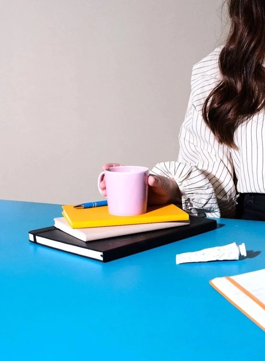 En person håller en rosa mugg i handen, placerad på tre staplade anteckningsböcker på en blå bordsyta.