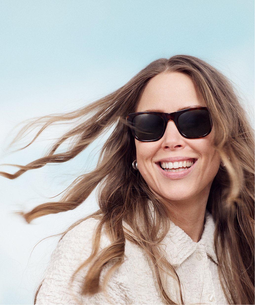 En glad kvinna med lockigt hår, solglasögon och vit jacka, som rör sig i vind.