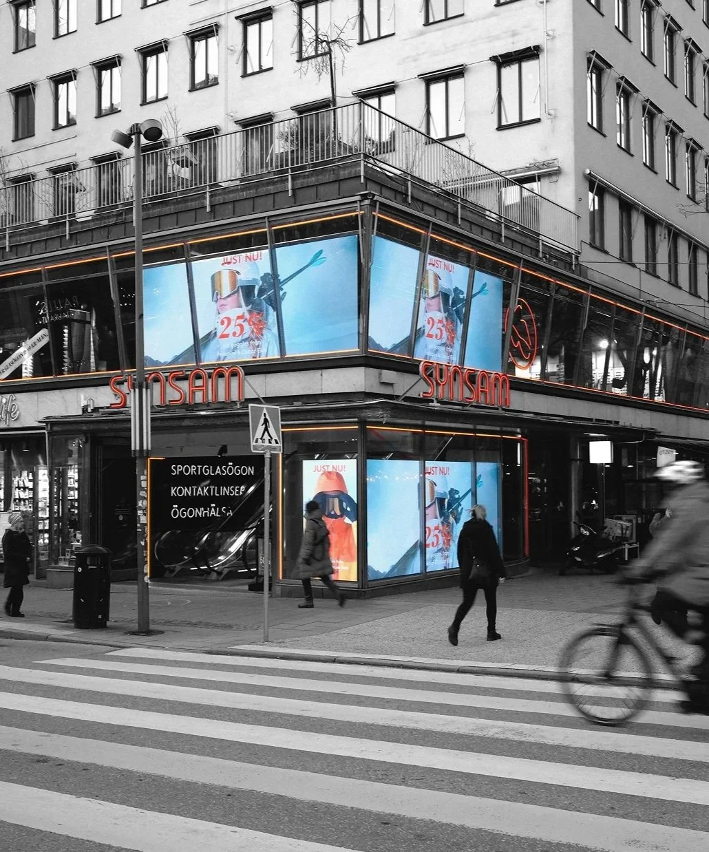 Bild av en gatukorsning med en byggnad som har två våningar och stora digitala reklamskyltar. Skyltarna visar en reklam för skid- eller snösport, med en person i skidutrustning och texten 'JUST NU!'. Gatan har en zebraövergång och några människor går och cyklar förbi.