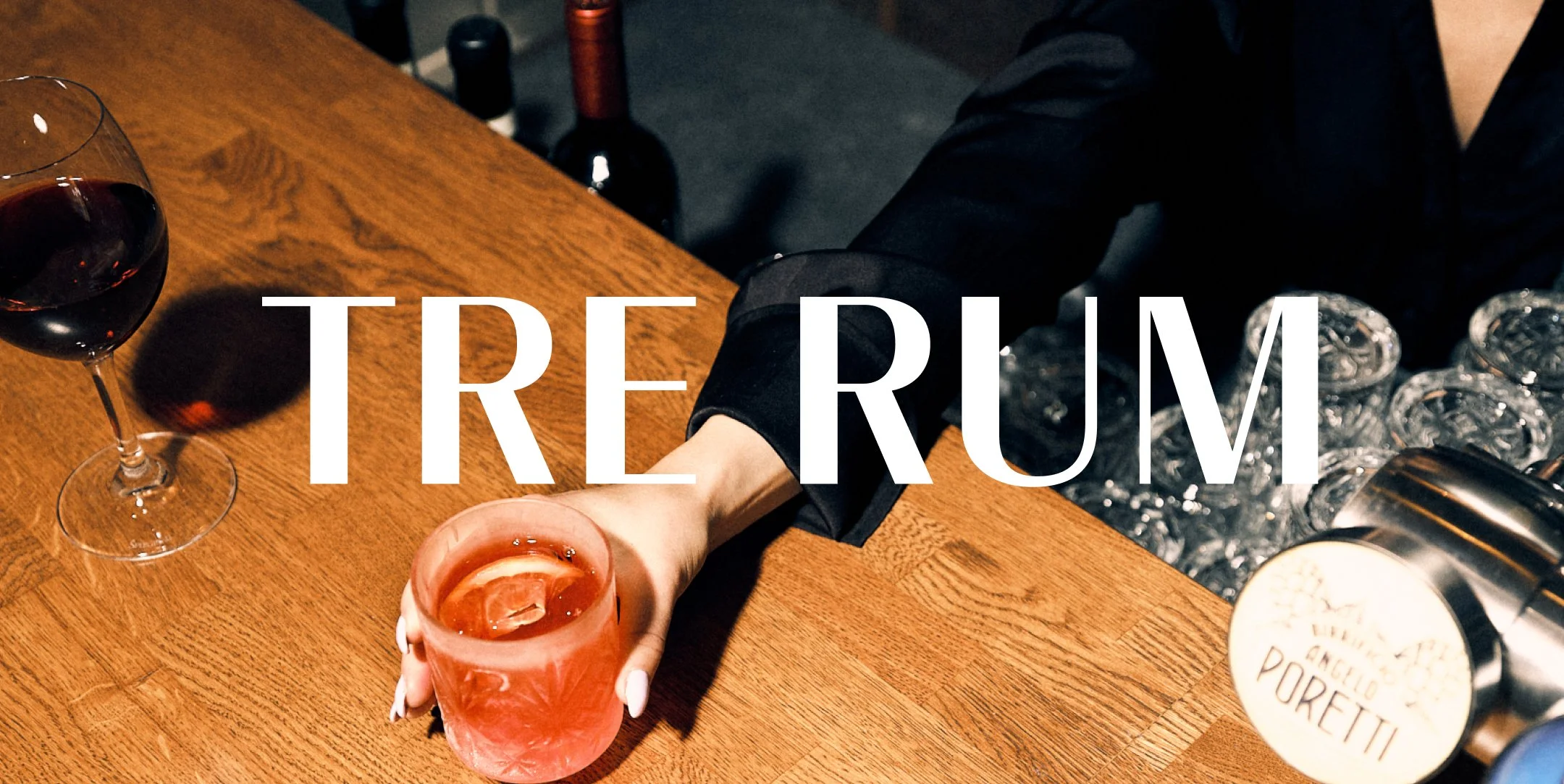 En person som serverar en rosa drink med citron på ett barbord av trä, med ett glas vin till vänster och flaskor i bakgrunden. Texten 'TRE RUM' över bilden.