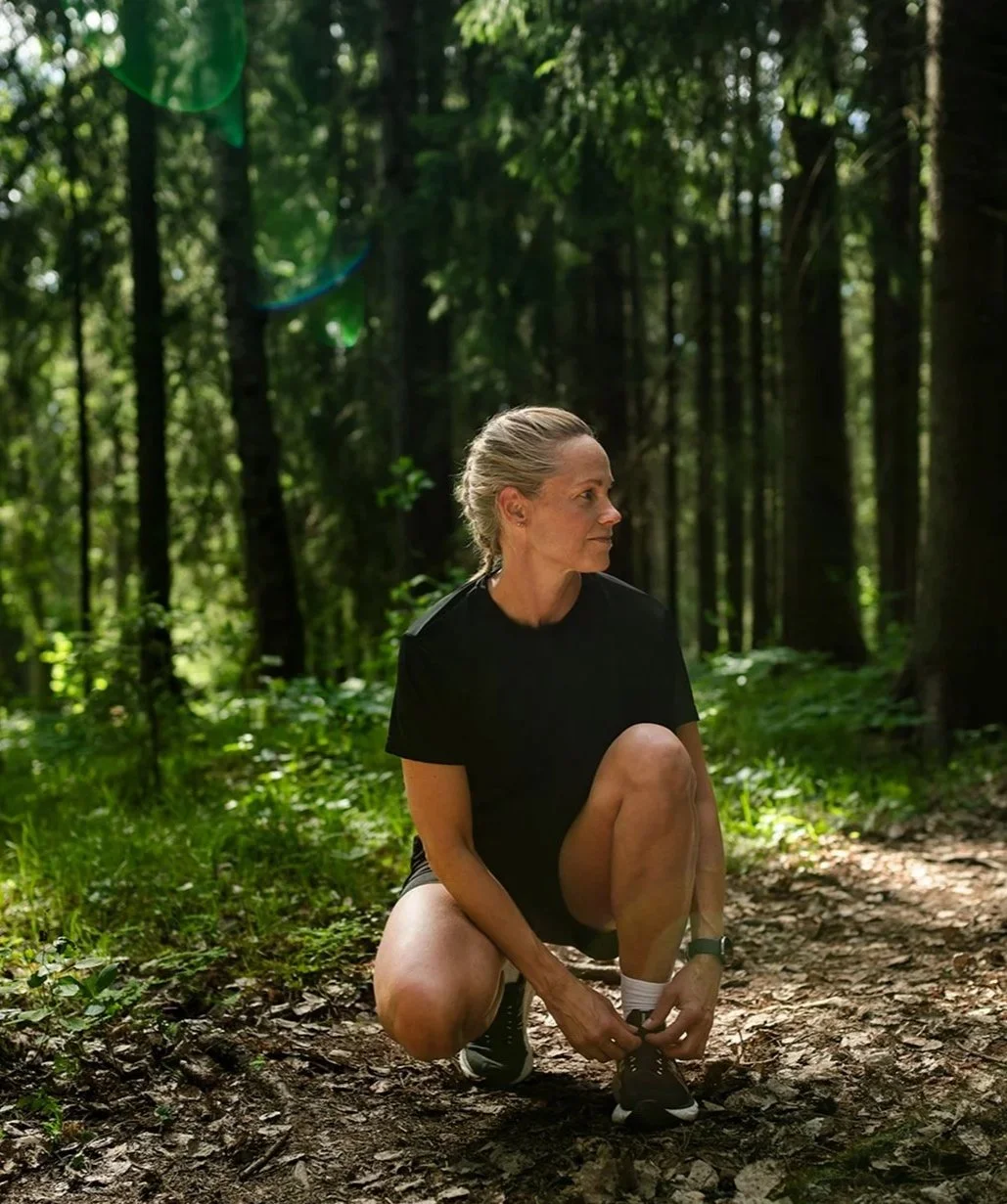 En äldre kvinna som knäpper sina skor i en skog, omgiven av grönt lövverk och träd.