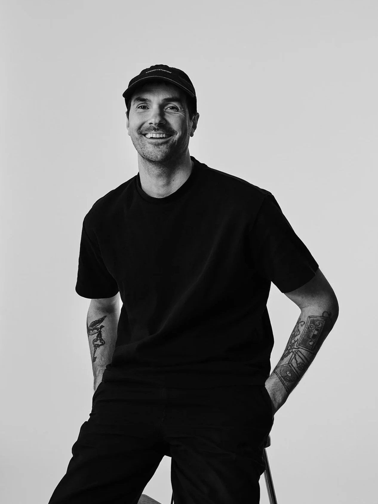 Ung man med tatueringar på armarna, svart tröja och svart keps, ler mot kameran, sitter på en stol mot en ljus bakgrund.