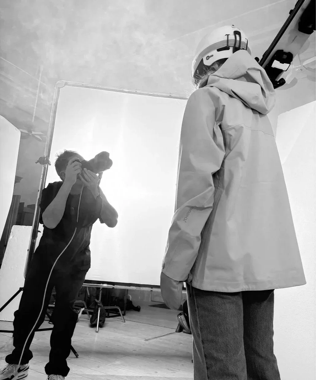 En person tar en självbild i spegeln, klädd i en vit jacka och hjälm, medan en fotograf tar bilden.