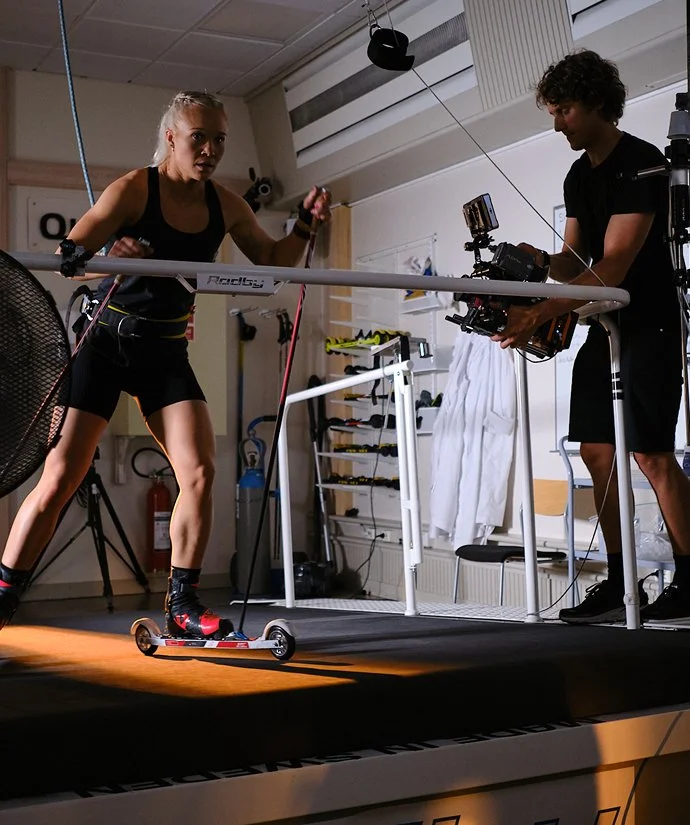 Kvinna testar motionscykel på filmuppsättningen medan en man filmas med professionell kamera.