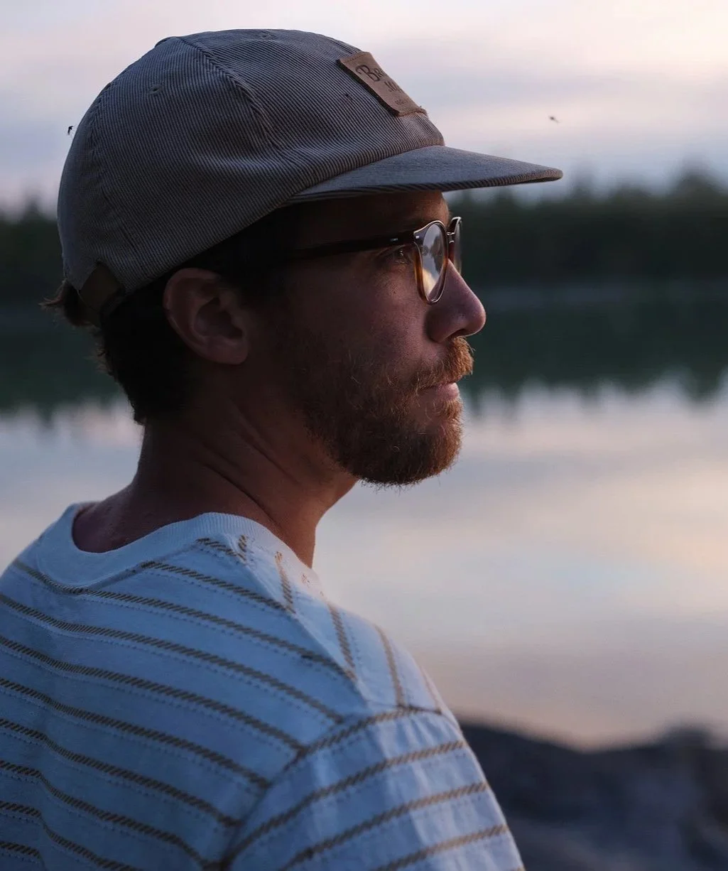 Man med glasögon och mössa tittar ut över vattnet vid solnedgången.