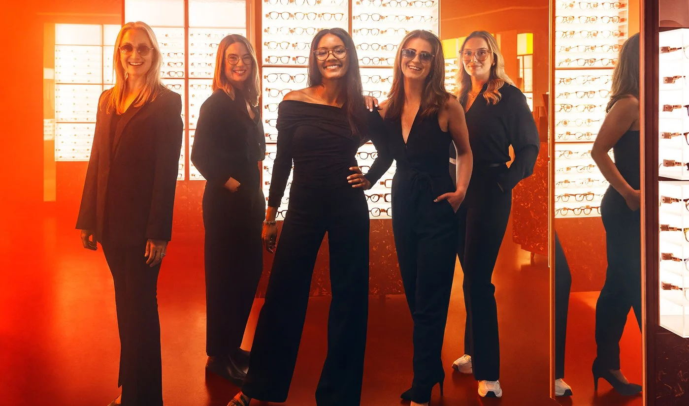 Fem kvinnor framför glasögonvägg i en optikerbutik, alla bär mörka kläder och visar leenden.