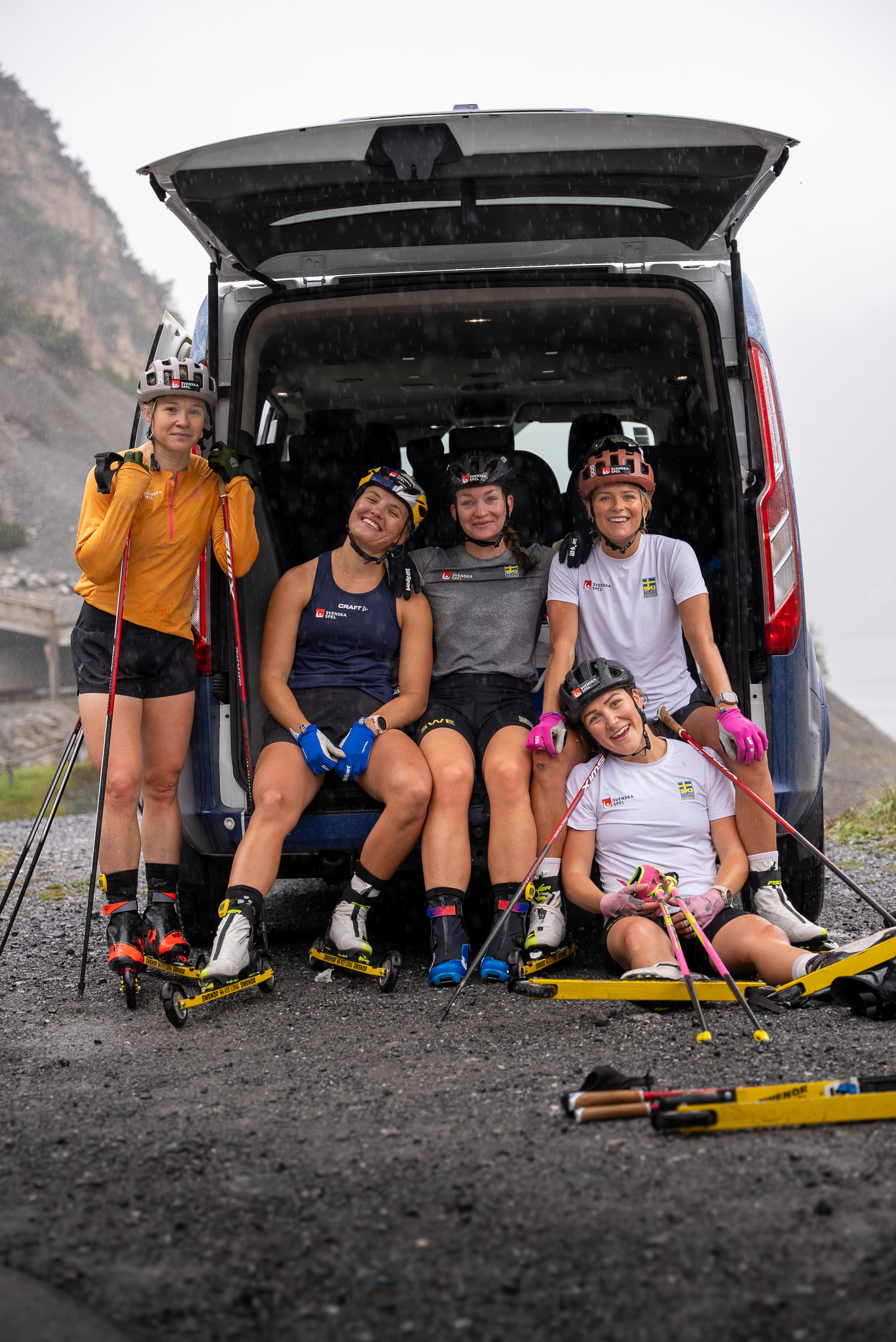 Fem kvinnor i skidutrustning, some allvar och glädje, sitter och står framför en öppen skåpbil på en regnig dag, i ett bergsområde.