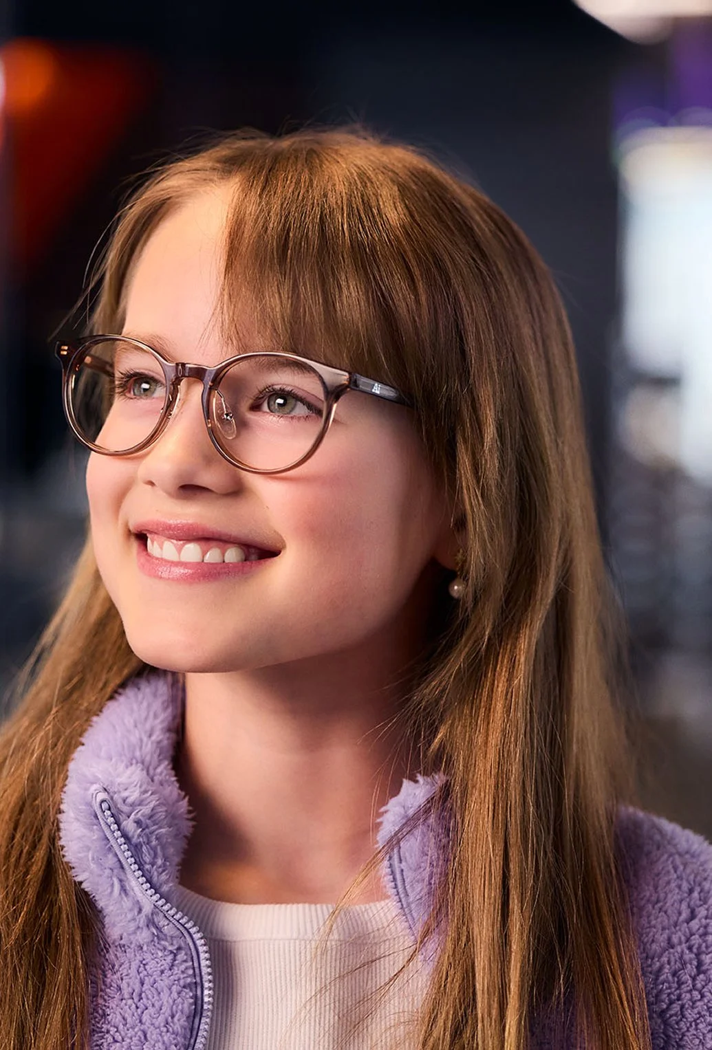 Ung flicka med brunt långt hår, glasögon och ljuslila fleecejacka, ler och tittar åt sidan i en inomhusmiljö.