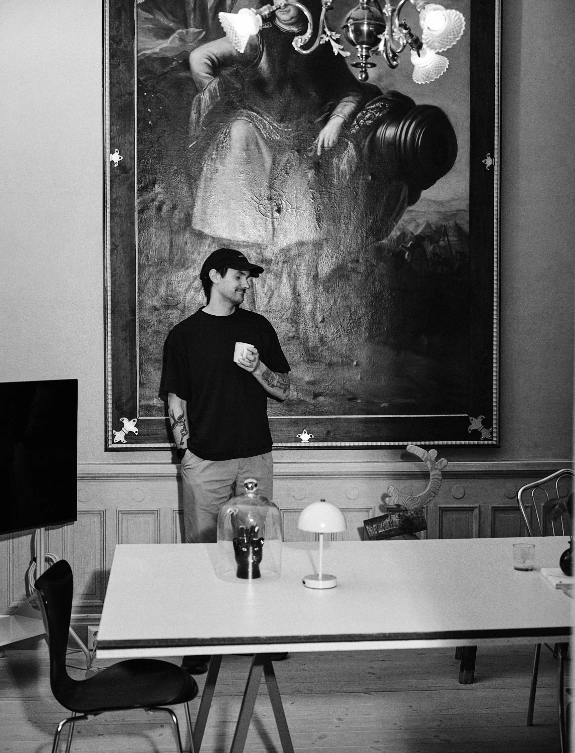 En ung man står i ett rum med ett stort tavla på väggen bakom honom. Han håller en kaffekopp och tittar åt sidan. På bordet framför honom finns en liten lampa och en glasburk med en svart katts huvud.