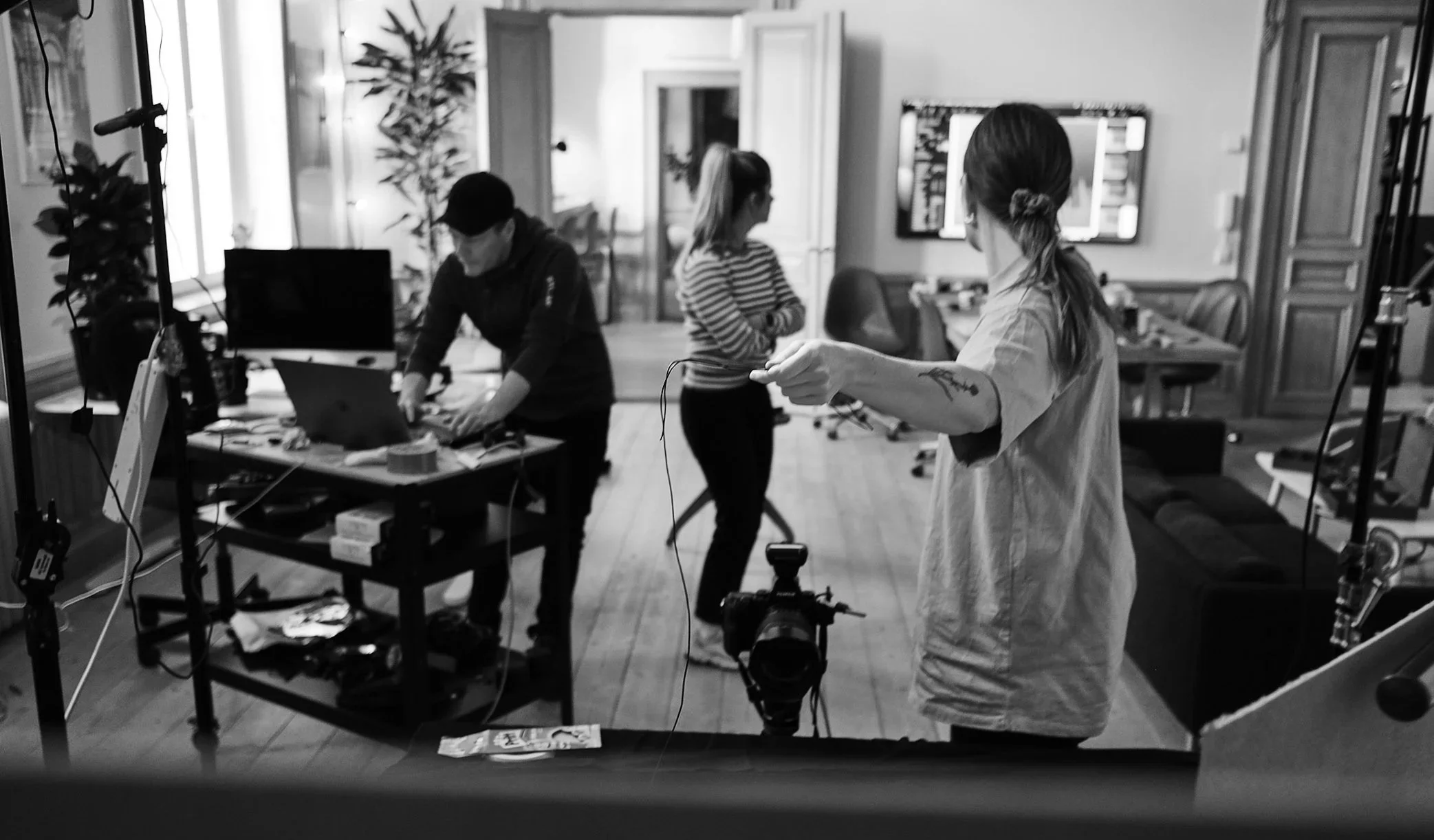 Tre personer i ett hem, där en person är vid ett bord med elektronik och två andra står, en med armar korsade och en annan som håller ett objekt.