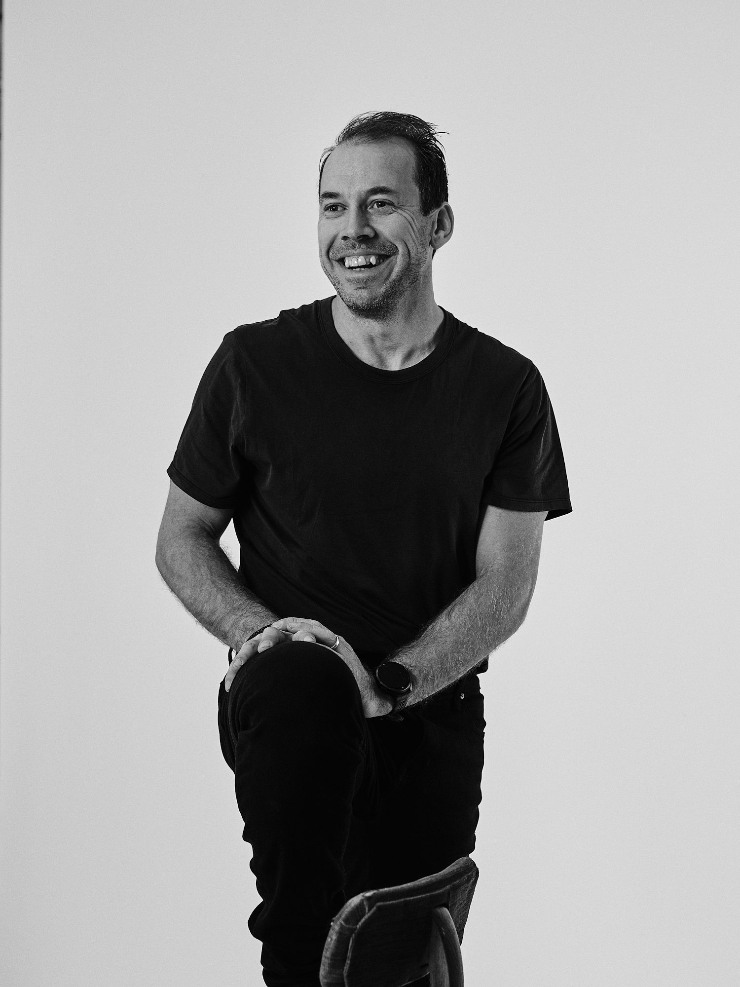 En man som ler och sitter på en stol, klädd i svart T-shirt och svarta byxor, med ett svart klocka på armen, mot en enkel ljus bakgrund.