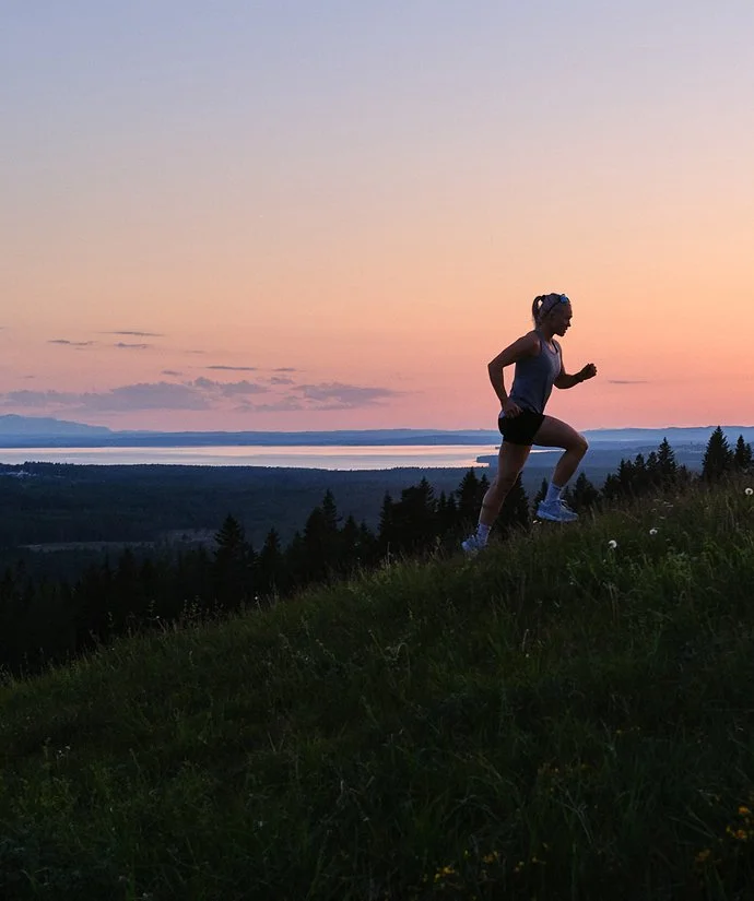 En kvinna löptränar uppför ett grönskande kullrike under en vacker solnedgång.