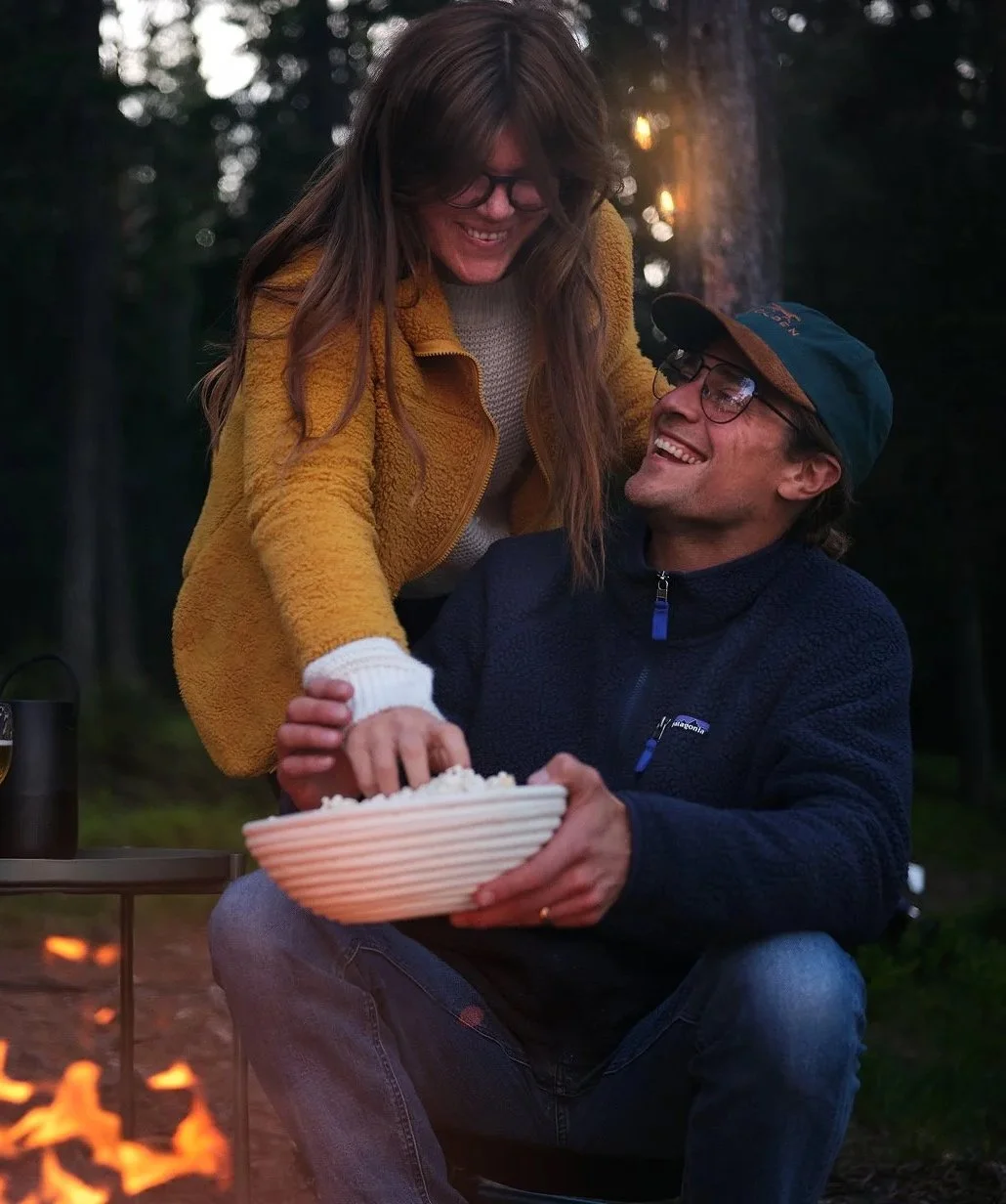 Två personer, en man och en kvinna, sitter utomhus i skogen, ler och delar ett skål med mat, framför en liten eld.