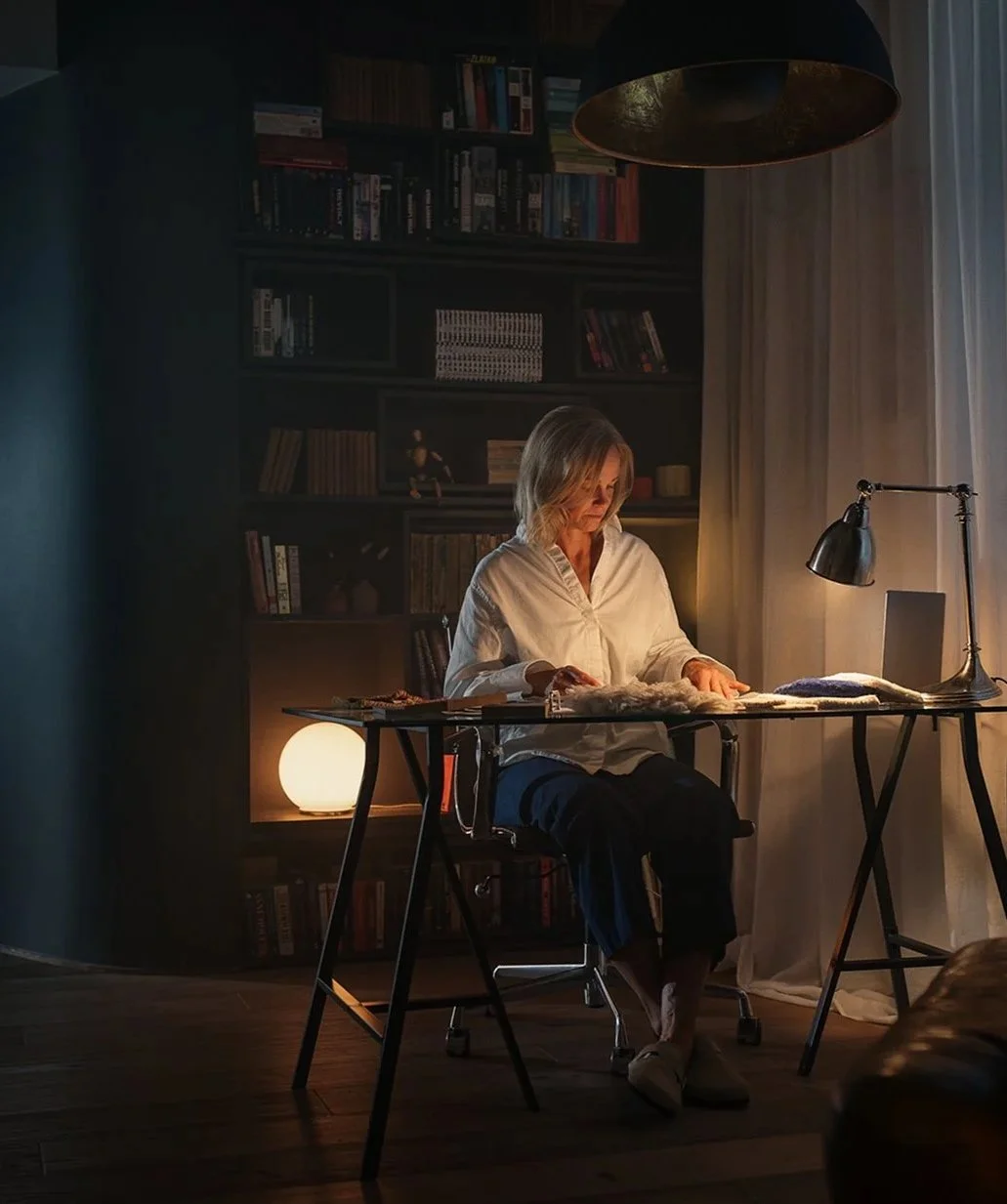 Kvinna som sitter vid ett skrivbord i ett mörkt rum, läser eller skriver, med en ett litet ljus, en skrivbordslampa och böcker i bakgrunden.