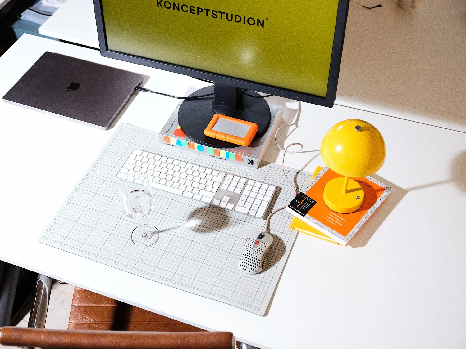 En vit arbetsyta med ett datorskärm, MacBook, trådlös keyboard, mus, glasvatten, ljusgul bordslampa, ett ex antal böcker, bortskymt stöd för laptop, och ett glas. Det finns även ett litet orange föremål under skärmen.