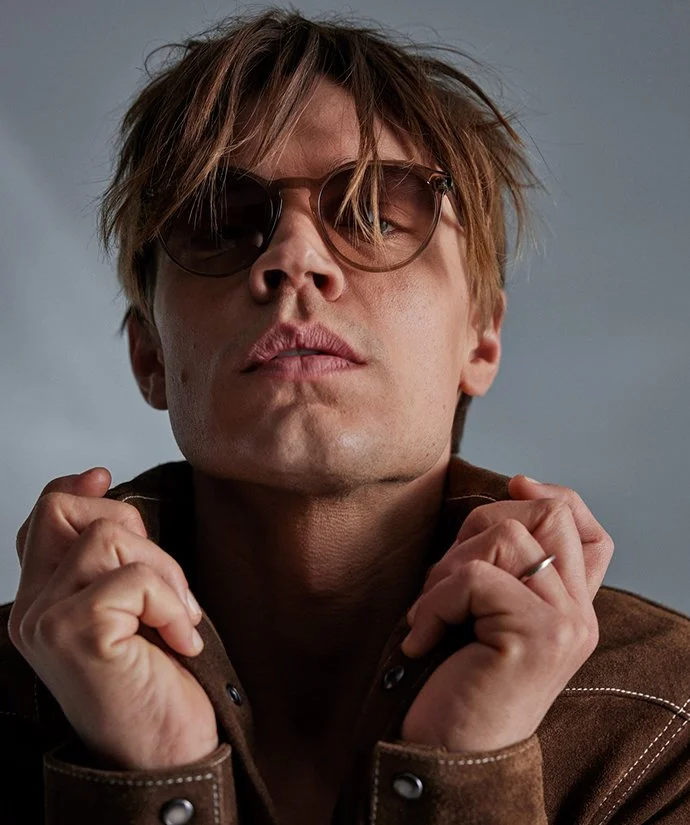 Ung man med solglasögon tar av sig en brun läderjacka.