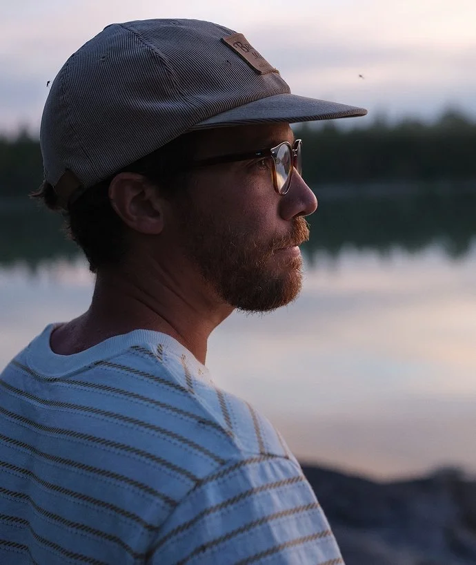 En man med glasögon och skägg, som bär en ljusblå och vit randig tröja och en grå keps, blickar mot vattnet under solnedgången.