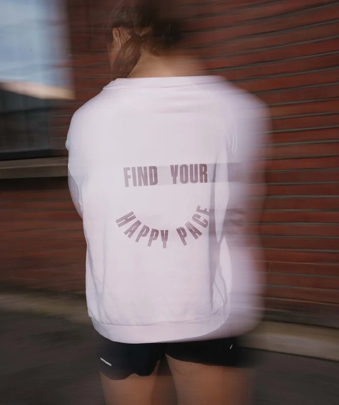 En person i vit tröja och svarta shorts står framför en röd tegelvägg, med texten "FIND YOUR HAPPY PACE" på ryggen.