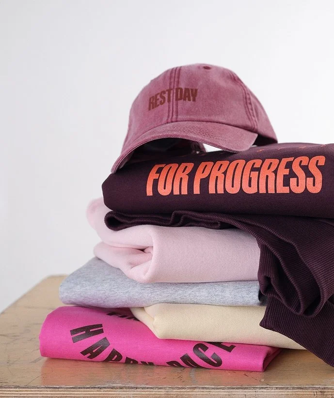 En hopfällbar ställning med en grå keps med texten 'REST DAY', ett svart plagg med texten 'FOR PROGRESS', ett ljusrosa plagg, ett grått plagg, ett gult plagg och ett rosa plagg med svart text.