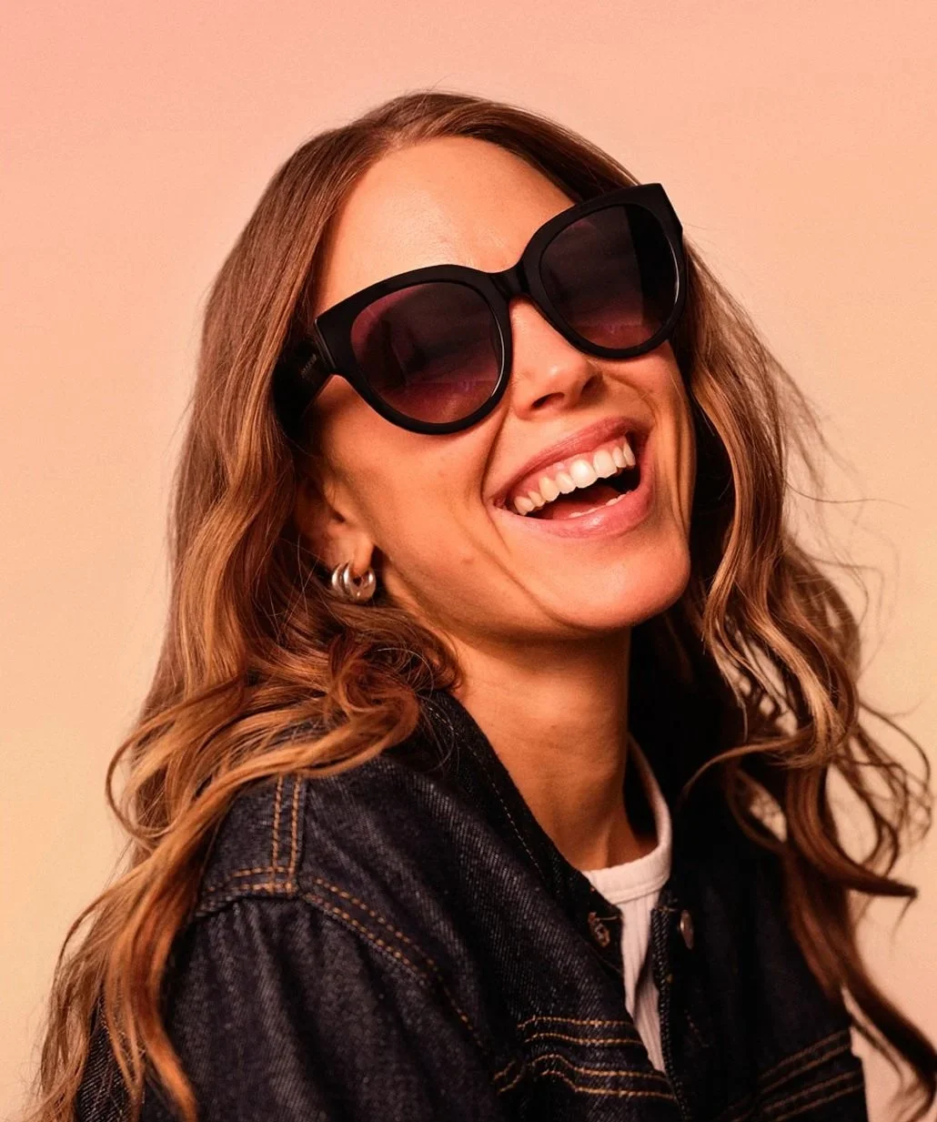 En leende kvinna med lockigt brunt hår, svart solglasögon och denimjacka mot en beige bakgrund.