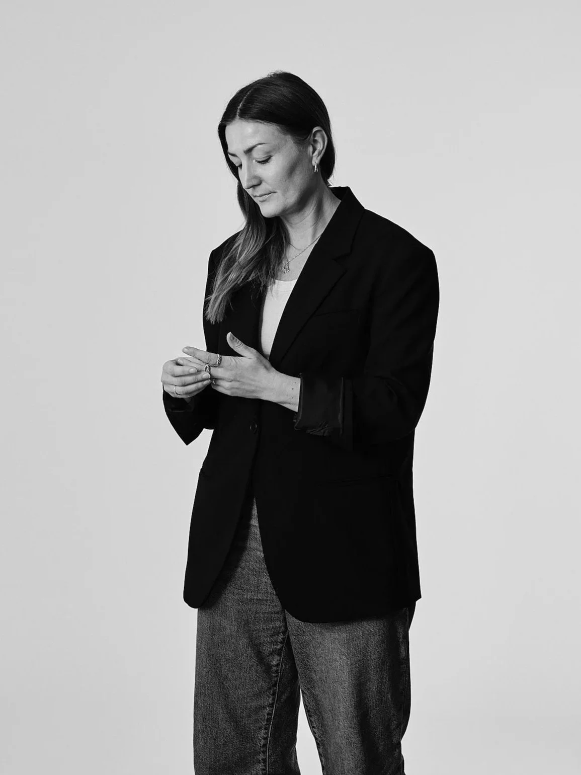 En kvinna i svart kavaj och jeans står mot en ljus bakgrund, tittar ner och justerar ett ring på sitt finger.