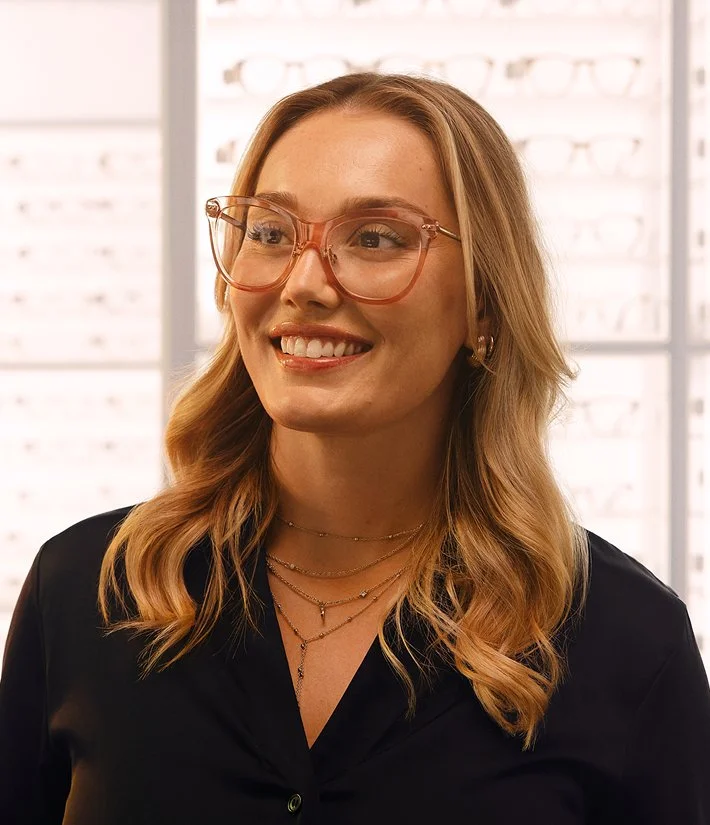 En glad kvinna med halvlång lockigt hår, glasögon och smycken i en optikerbutik.