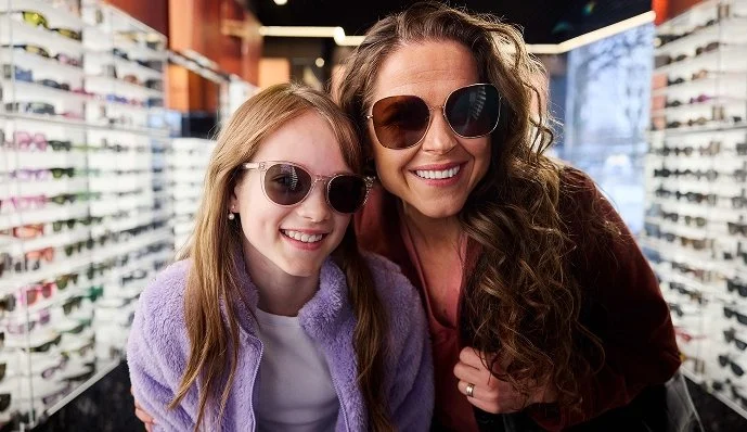 Två kvinnor med solglasögon i en optikbutik, glada och smilande.