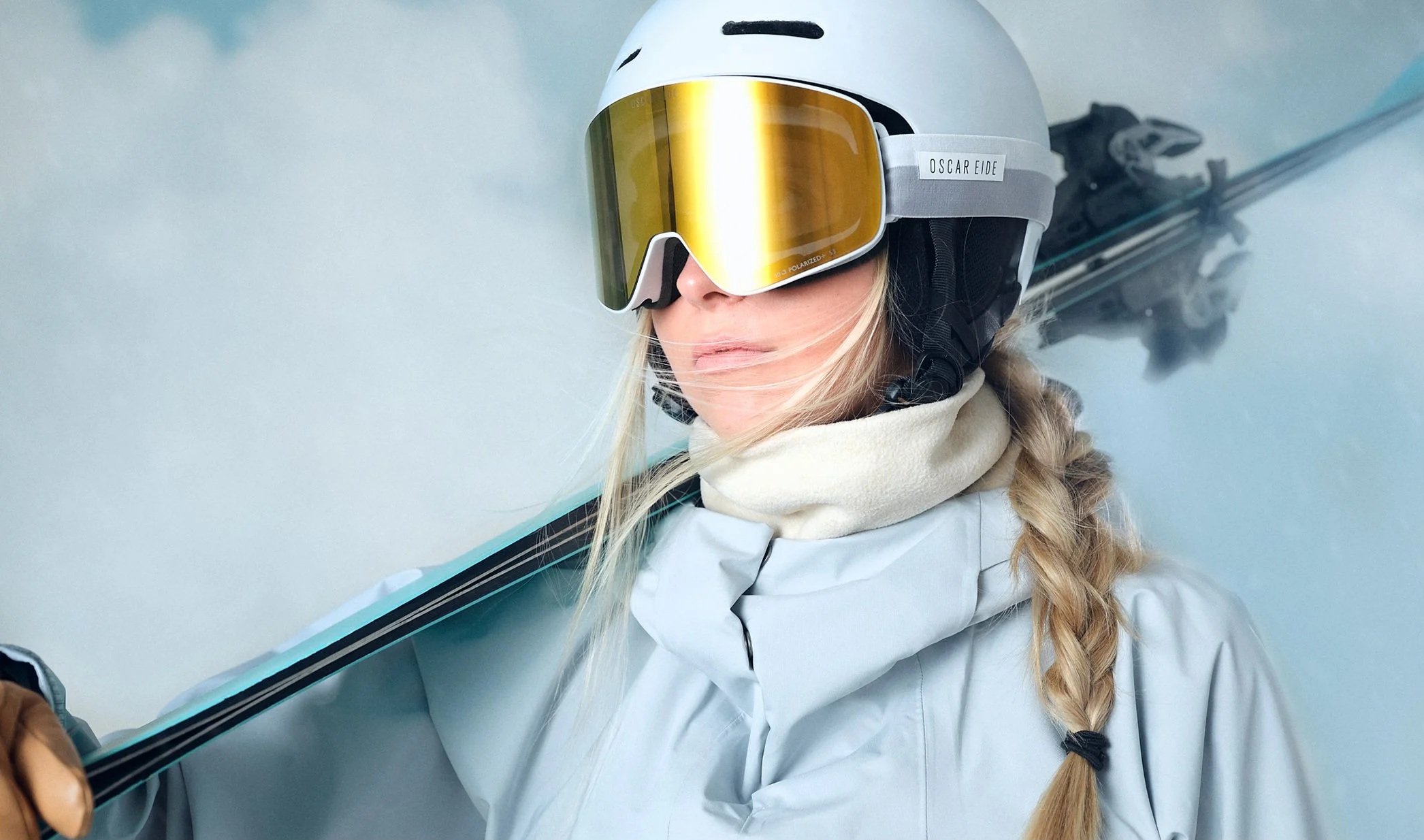 En kvinna med skidutrustning, inklusive hjälm och skidglasögon, står i snön och håller ett par skidor över axeln.