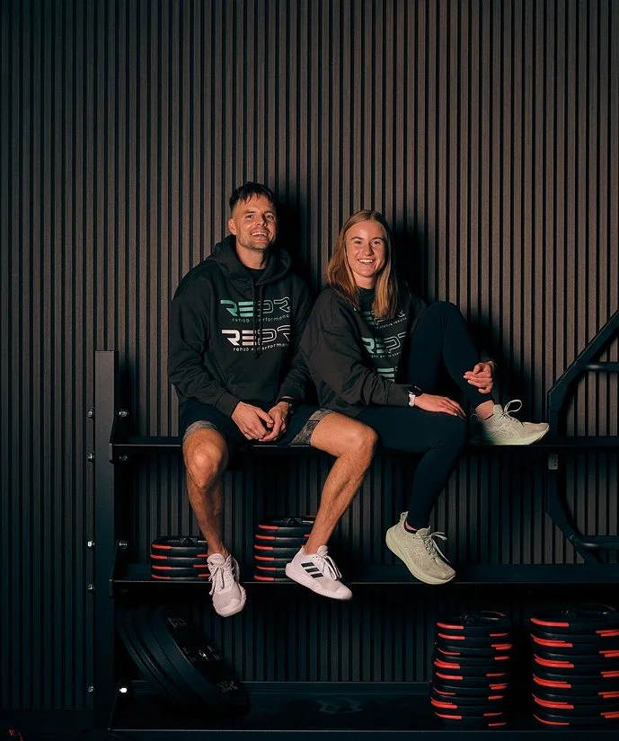 Två personer sitter på en svart träningsbänk, båda bär svarta hoodie och träningsshorts, och ler mot kameran. Bakgrunden är en svart vägg med vertikala ragla.