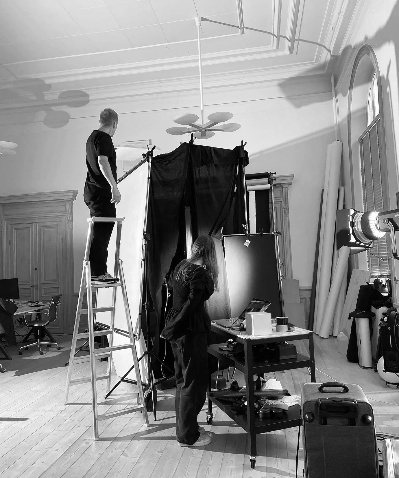 Två personer som arbetar med en fotostudio, en människa står på en stege och justerar en backdrop, den andra stående framför en belysningsutrustning.