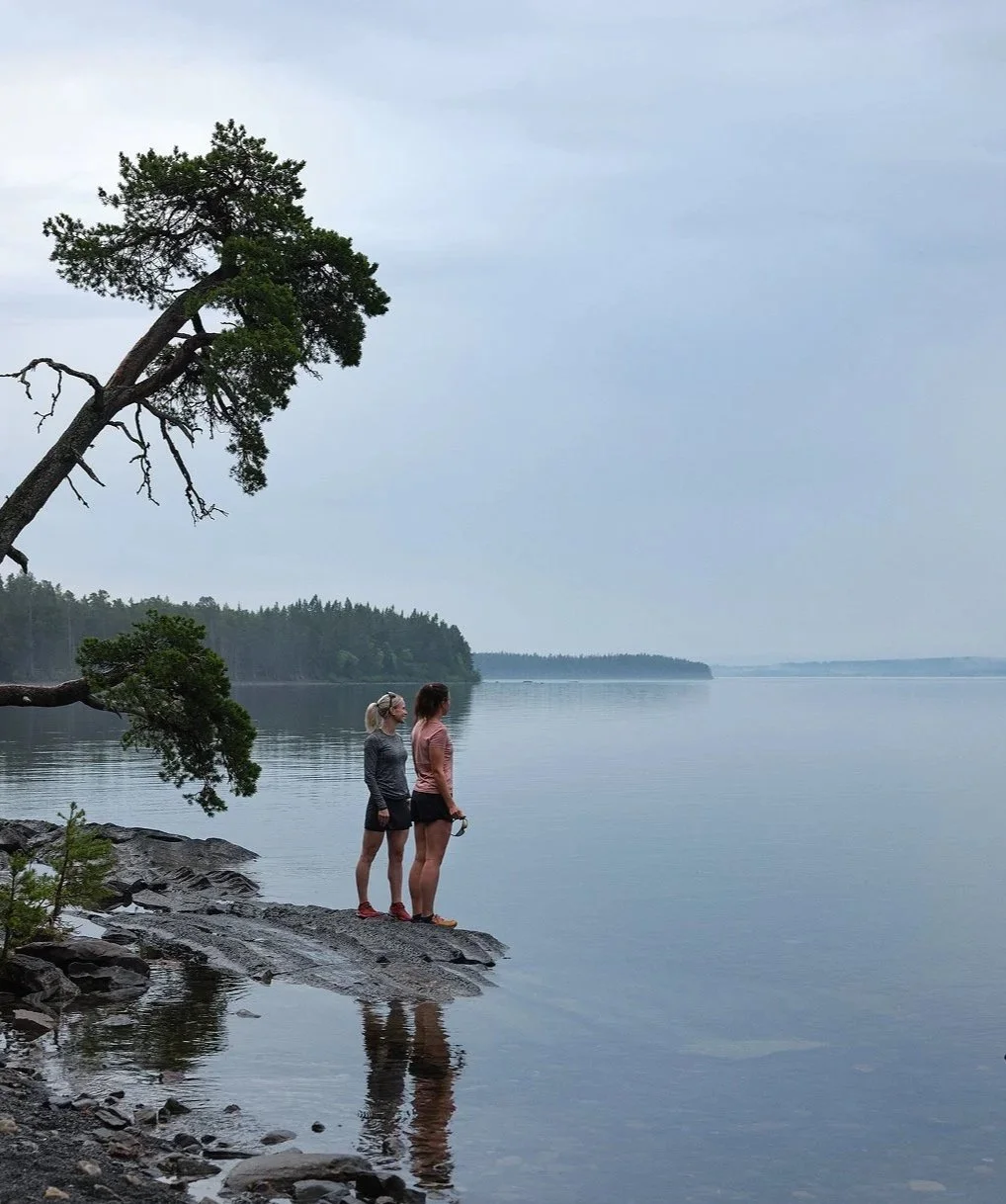 Två kvinnor står på en sten vid en sjökant, tittar ut över vattnet, med en stor trädgren som hänger över dem, omgivning av skog och lugnt vatten.