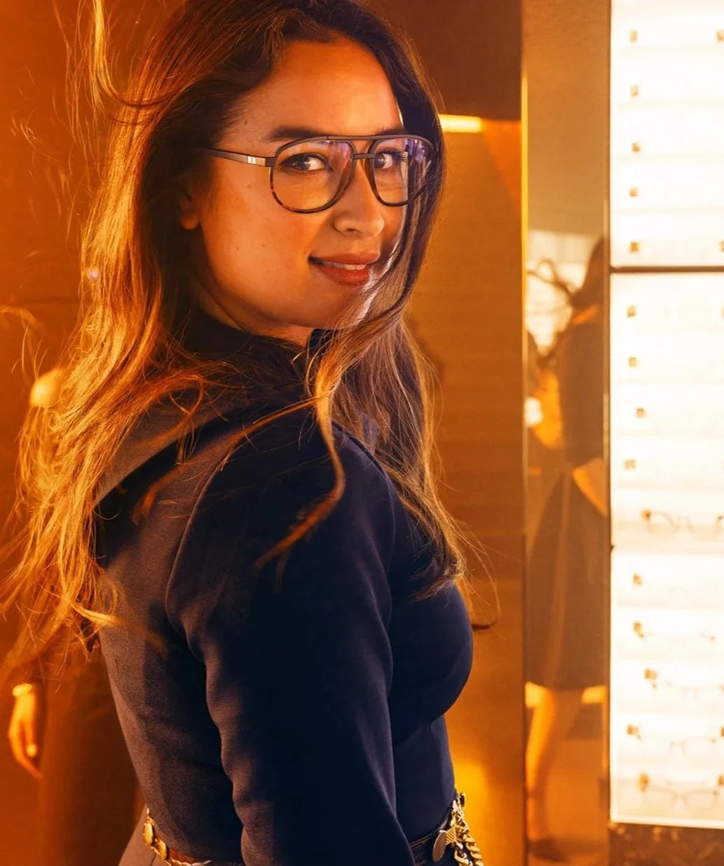 En kvinna med glasögon som ler, med långt lockigt hår, står i ett varmt, gult ljus, troligtvis i en butik eller ett bibliotek.