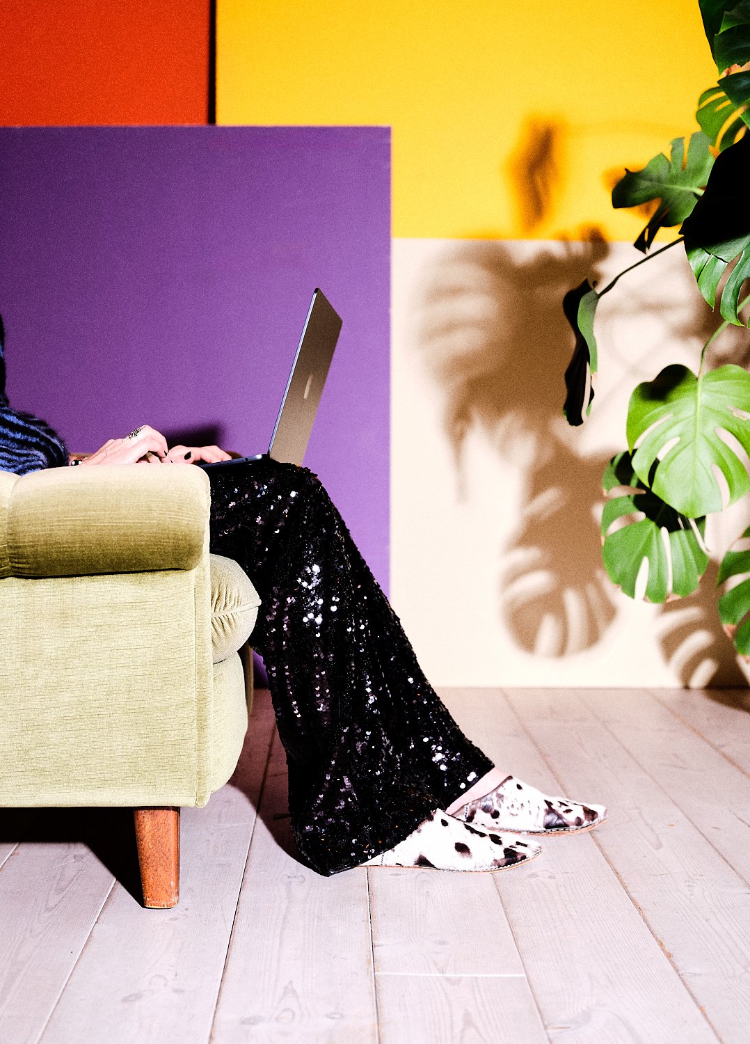 Person som sitter på en gul fåtölj och använder en laptop, med färgglada väggar och en växt i bakgrunden.