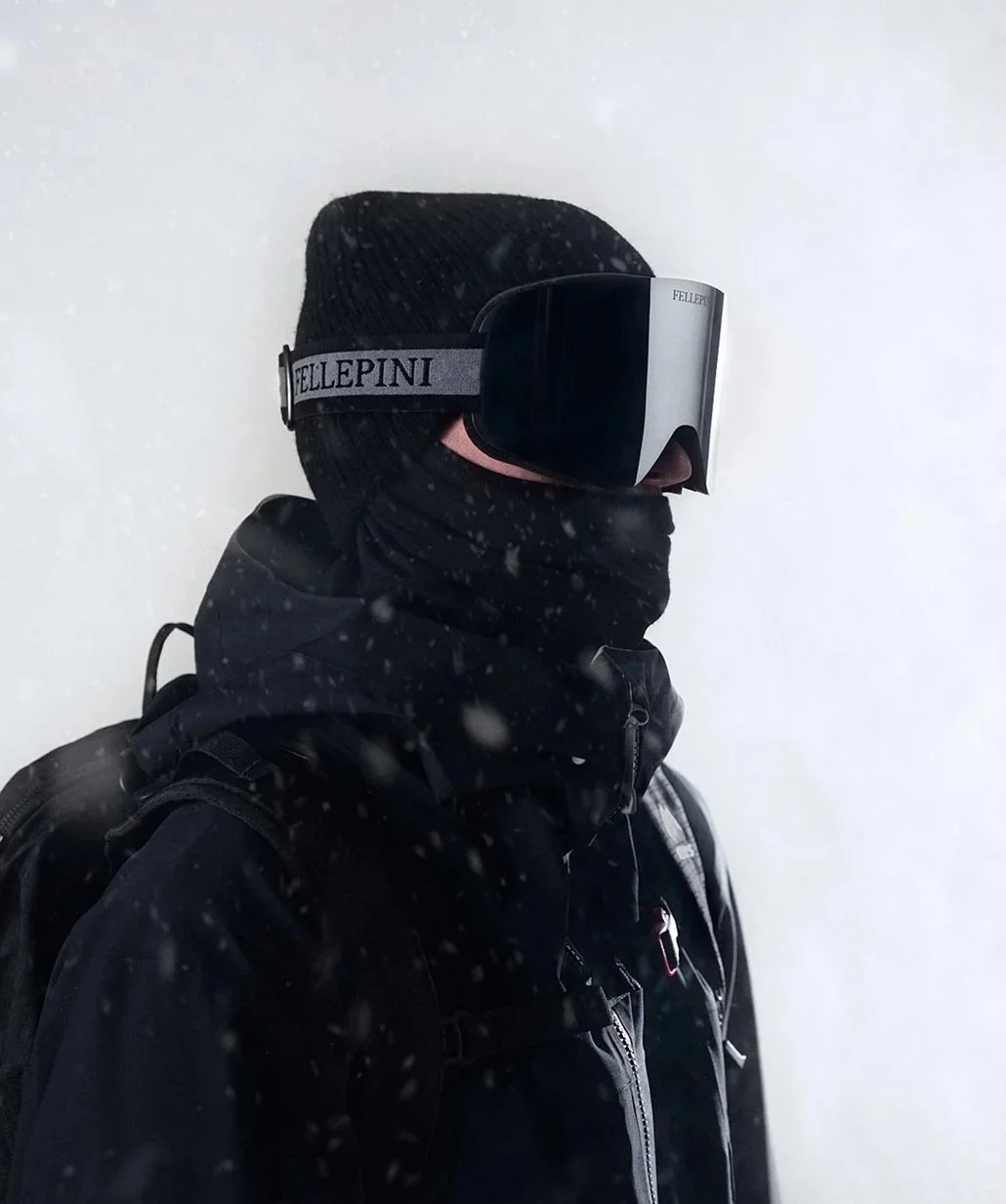 Person iförd skidutrustning, inklusive svart mössa, skidglasögon och svart jacka, stående mot en vit snöig bakgrund.
