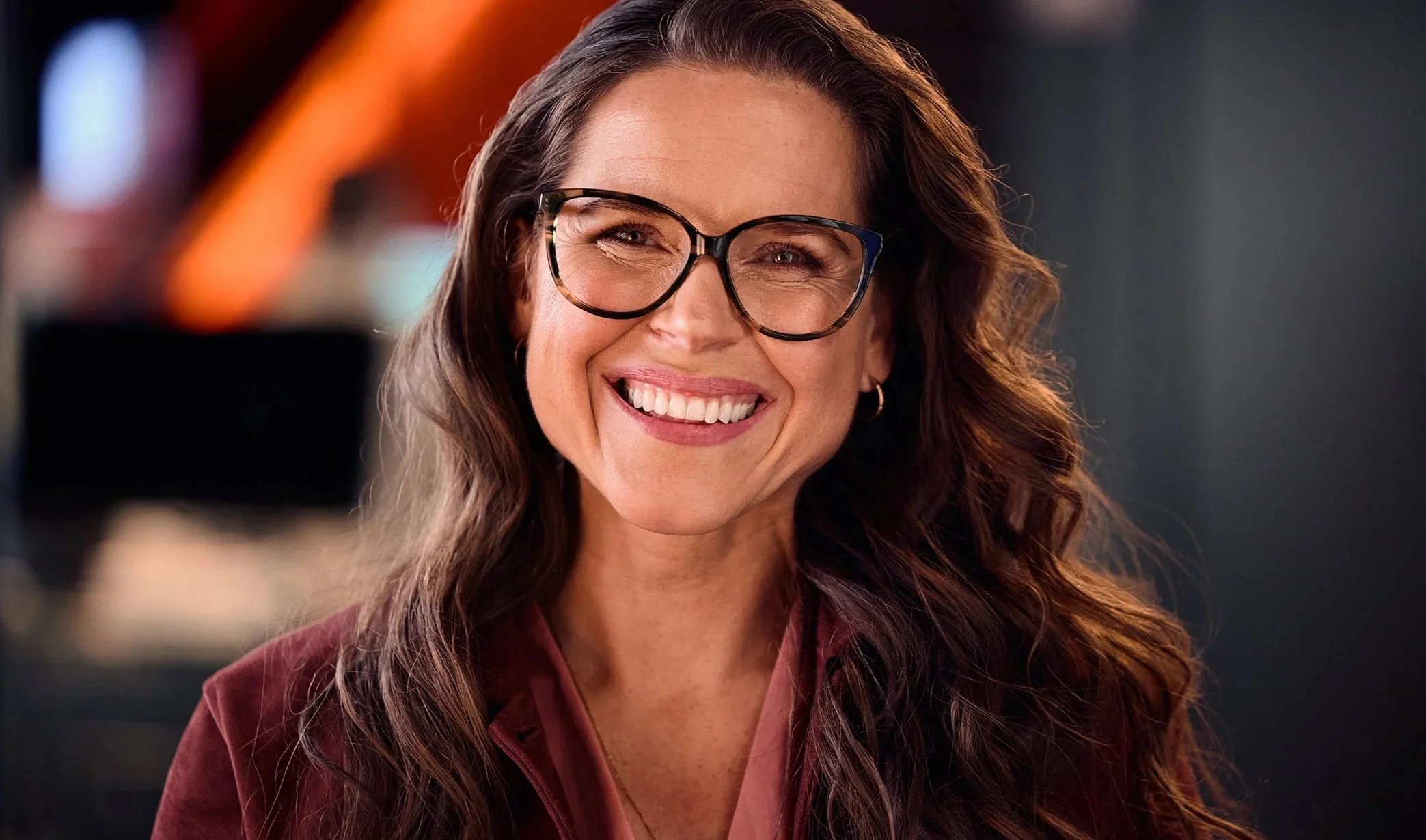 En leende kvinna med glasögon, långt lockigt hår, iklädd en mörkröd jacka och rosa blus, i en modern inomhusmiljö.