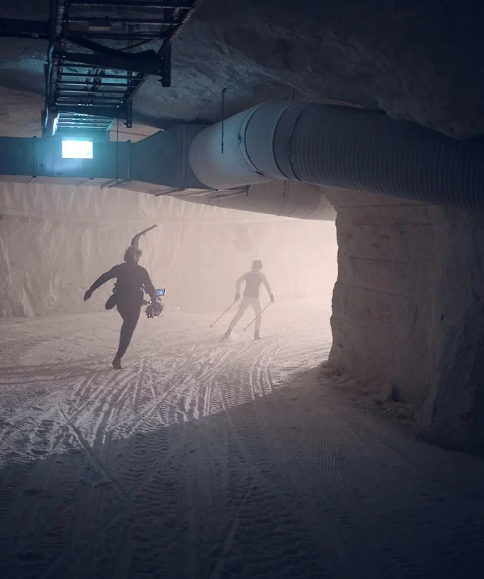 Två personer som åker skidor genom en snöig underjordisk grotta eller tunnel, med en stor ventilationsrör och ett litet fönster eller ljus bakom dem.