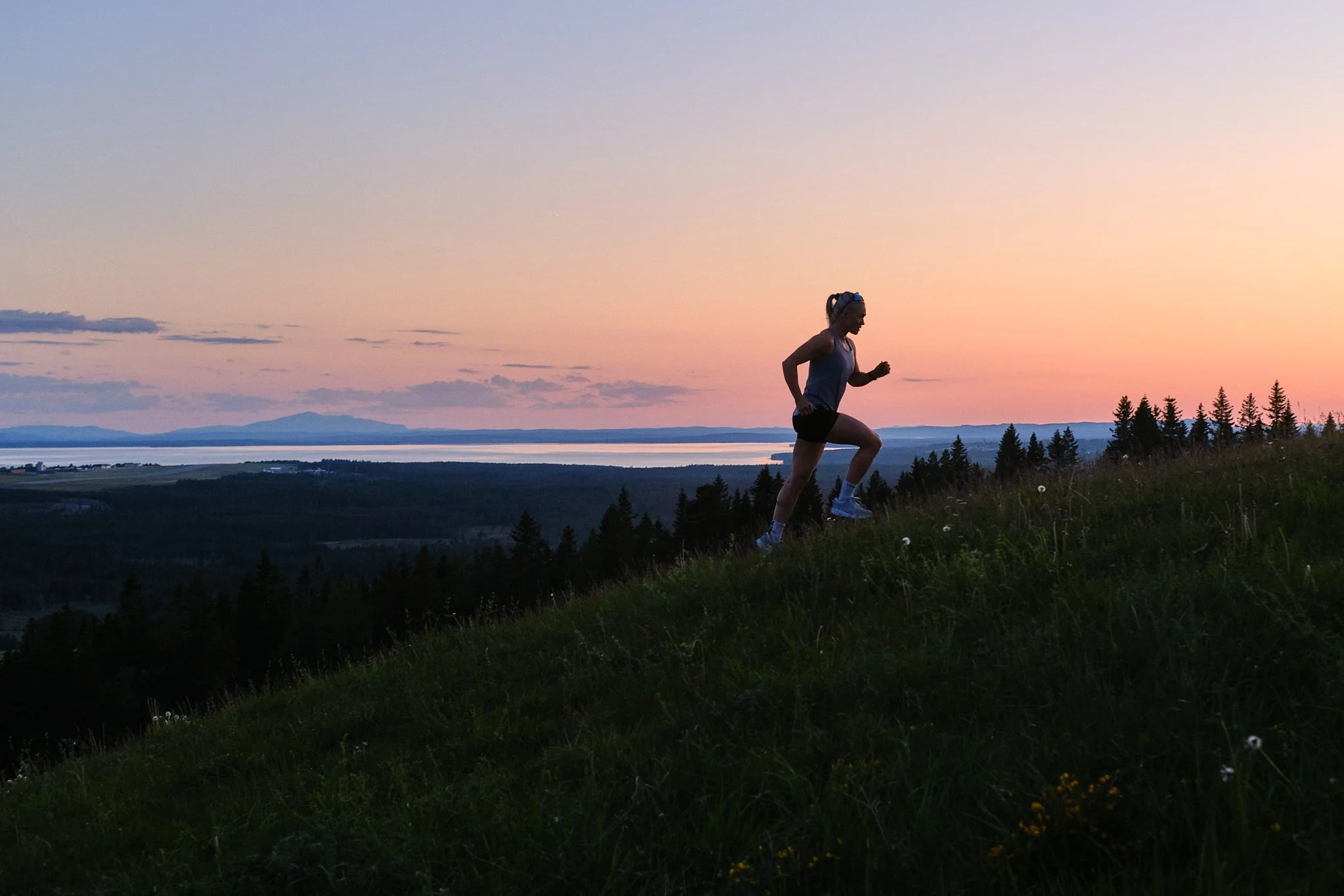 En kvinna springer uppför en gräsbeklädd backe vid solnedgången, med en sjö och träd i bakgrunden