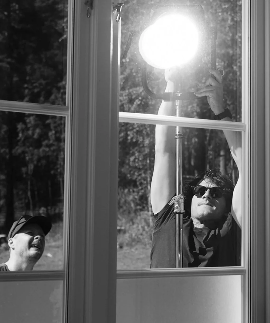 Två män utanför ett fönster, en av dem tar ner kläppen, den andre tittar på och har på sig solglasögon, i svartvitt.