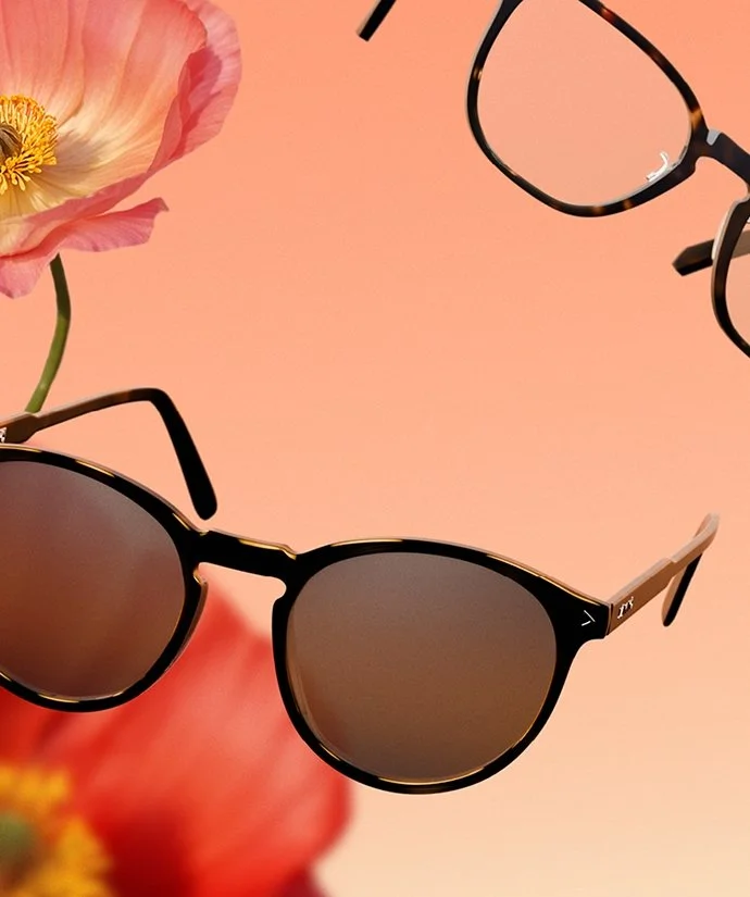 Två par solglasögon, ett par i svart med bruna linser och ett annat i av tortoise-mönster, ligger på en rosa bakgrund med blommor i bakgrunden.